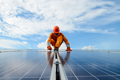 Image shows a man wearing an orange hardhat and uniform working on a solar power station. 